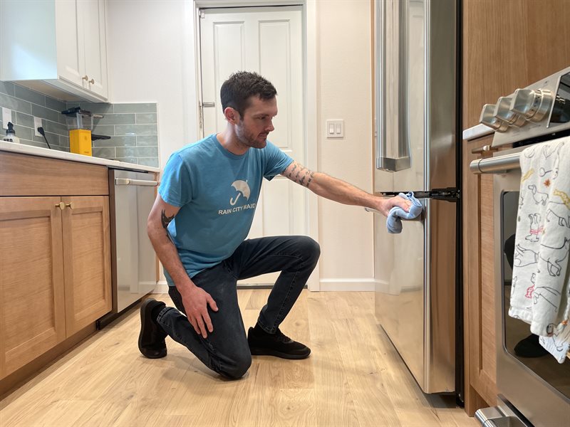 A Rain City Maids cleaner kneeling to wipe a lower kitchen cabinet