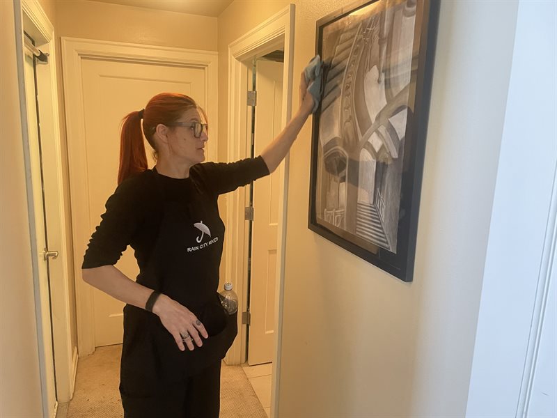 A Rain City Maids cleaner dusting a framed picture in a hallway